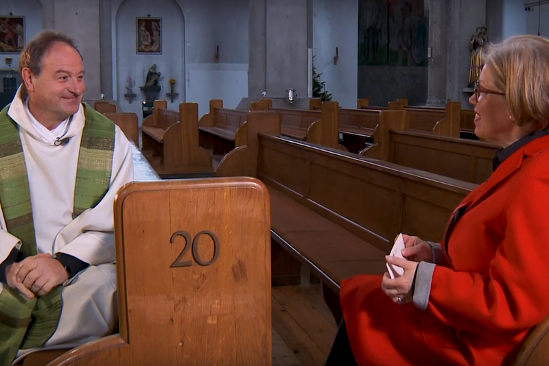 Pfarrer Rainer Maria Schießler und Andrea Haagn sitzen in Kirchenbänken und unterhalten sich. 