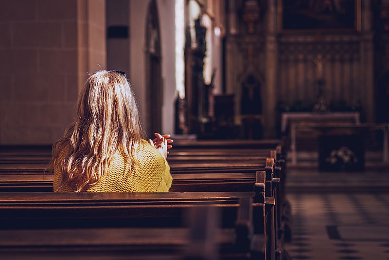 Eine Frau sitzt in einer hinteren Kirchenbank in einer leeren Kirche, blickt zum Altar und betet.