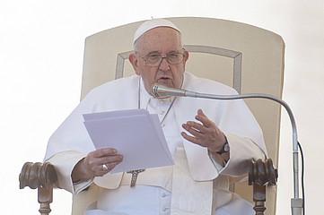 Papst Franziskus sitzt auf einem Stuhl und hält ein Manuskript in der Hand 