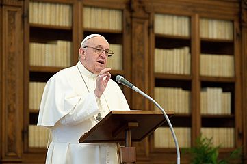 Papst Franziskus steht am Ambo und predigt. Vor ihm steht ein Mikrofon am Stativ, hinter ihm sind große Schränke mit Büchern zu sehen. 