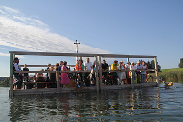 Viele Menschen verschiedenen Alters sitzen gemeinsam auf einem Floß. Mit Hintergrund ein großes Kreuz, dazu der See.