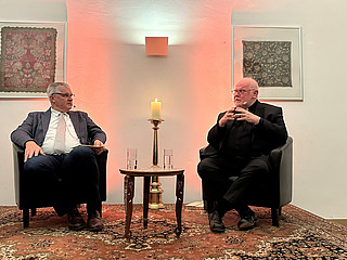 Zwei weiße, ältere Männer sitzen auf Sesseln in Talksetting und sprechen miteinander.