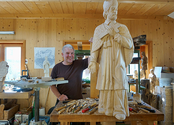 Christian Dögerl steht in seiner Werkstatt hinter der Werkbank. Darauf steht die noch nicht ganz fertige Nepomuk-Figur, drumherum liegen Werkzeuge und Schnitzeisen.