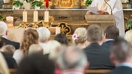 Ein Priester steht an einem festlich geschmückten Altar am Ambo, in der Hand hält er ein Manuskript. Die Kirche ist gut gefüllt, das Gesicht des Priesters ist nicht zu sehen.