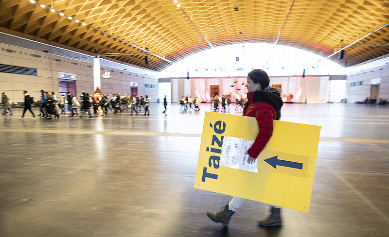 Frau trägt Schild mit Aufschrift "Taizé" in einer großen Halle, im Hintergrund Menschen
