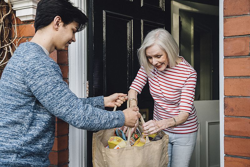 Junger Mann übergibt älterer Frau Einkäufe an der Haustür.