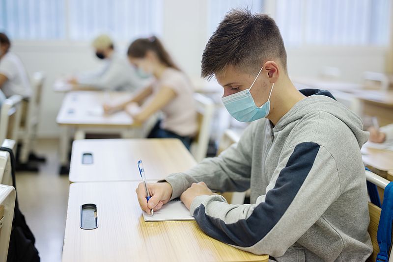 Mehrere Schüler sitzen mit einer Mund-Nase-Bedeckung und mit ausreichend Abstand in einem Klassenzimmer. Im Fokus ist ein Junge, der an seinem Platz sitzt und mit einem Stift etwas in ein Heft schreibt. 