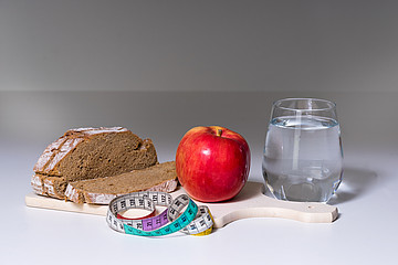 Auf einem Frühstücksbrett liegen Graubrot und ein Apfel, daneben steht ein Glas Wasser, davor liegt ein Maßband.