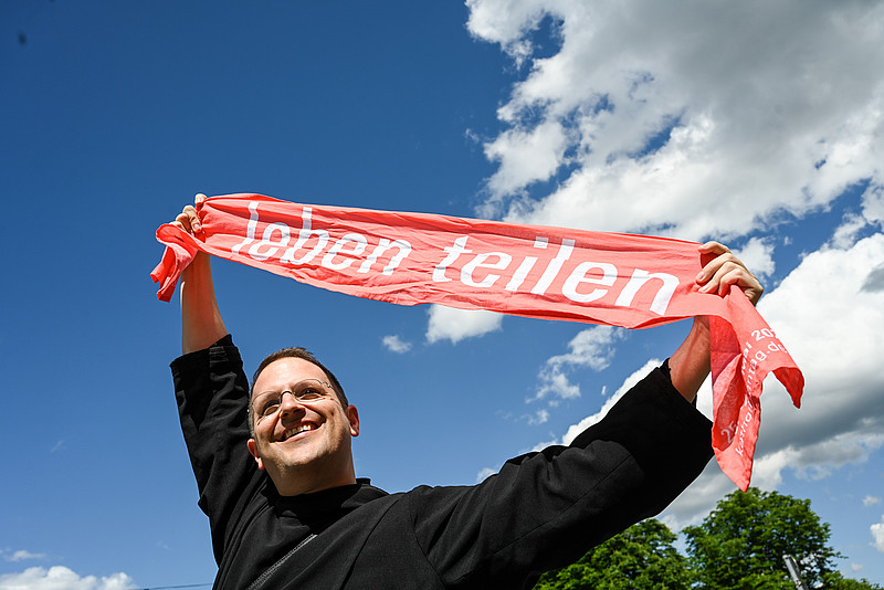 Ein lachender Mann hält einen roten Katholikentagsschal hoch über seinen Kopf mit der Aufschrift "leben teilen", dem Motto des 102. Deutschen Katholikentags, am 25. Mai 2022 in Stuttgart.