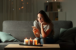 Frau sitzt auf Couch und schaut auf das Smartphone. Kerzen sind angezündet. 