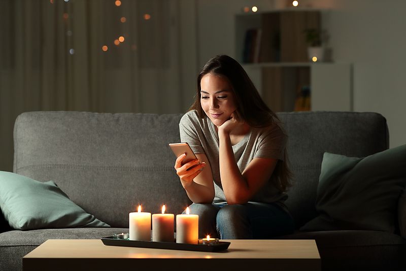 Frau sitzt auf Couch und schaut auf das Smartphone. Kerzen sind angezündet. 