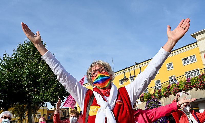 Eine Frau mit einem regenbogenfarbenen Mundschutz erhebt die Arme