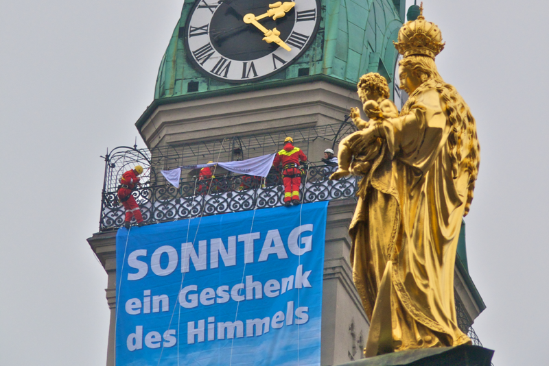 Plakat "Sonntag ein Geschenk des Himmels" auf dem Münchner Petersturm Marienstatue im Vordergrund