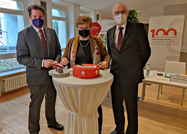 100. Geburtstag in Coronazeiten: Der Vorstand Thomas Schwarz (l.), Vorständin Gabriele Stark-Angermeier und Caritasdirektor Hermann Sollfrank stehen mit Maske an einem Tisch, auf dem die Geburtstagstorte steht.