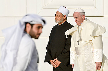 Zwei ältere Männer stehen nebeneinander, der rechte trägt ein weißes Gewand, ein Kreuz um den Hals und eine weiße Kopfbedeckung. Der Linke trägt ein schwarzes Gewand und eine andere weiße Kopfbedeckung. Im Vordergrund ist verschwommen ein Mann in weißem Gewand und mit einem weißen Kopftuch zu sehen. 
