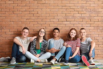 Fünf Jugendliche sitzen nahe zusammen an eine Mauer gelehnt auf dem Boden und umarmen sich. (Symbolbild) 