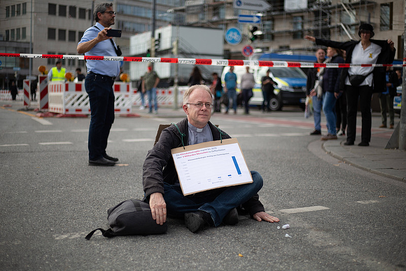 Jesuit Jörg Alt im Schneidersitz auf einer Straße mit einem Plakat vor sich
