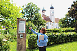 Dr. Petra Altmann an einer Impulstafel auf dem Benediktusweg mit dem Kloster im Hintergrund.