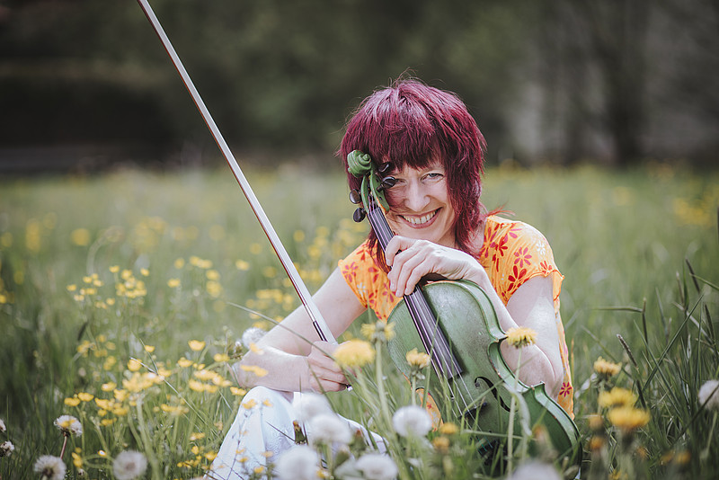 Monika Drosch in einer grünen Wiese sitzend mit ihrer ebenfalls grünen Geige.