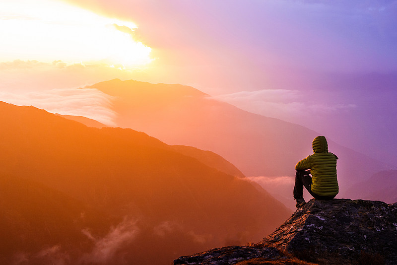 Person sitzt auf Felsen, umgeben von Bergen und schaut in den Sonnenuntergang