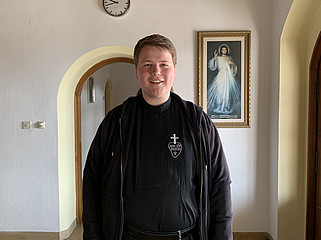 Frater Benedikt in schwarzem Gewand steht im Flur und lächelt.