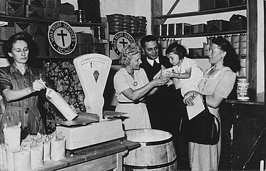 Schwarzweiß-Foto von der Essensausgabe der Caritas in der Heßstraße um 1946. Waren werden abgewogen und ein Kind auf dem Arm einer Frau greift nach einer Tüte, die ihm gereicht wird. In den Regalen im Hintergrund sind Waren gelagert.