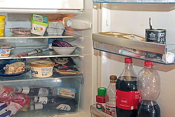 Überfüllter Kühlschrank, in dem sich u.a. eine große Flasche Cola, Wurst, Kuchen, Margarine, Bier, Eier und Joghurt stapeln.