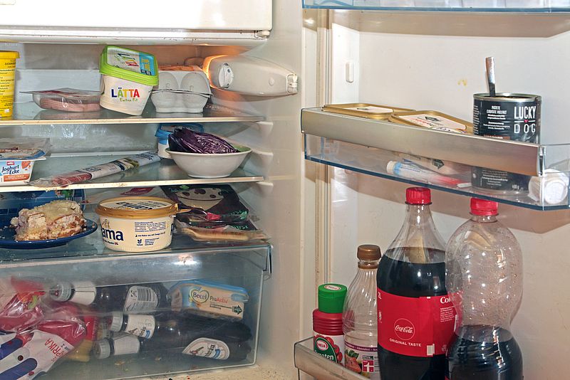 Überfüllter Kühlschrank, in dem sich u.a. eine große Flasche Cola, Wurst, Kuchen, Margarine, Bier, Eier und Joghurt stapeln.