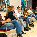 Junge Menschen sitzen in einer Reihe, eine Jugendliche liest von einem Blatt ab. 