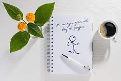 In der Mitte eines Tisches liegt ein aufgeschlagener Notizblock, indem steht "Ab heute gehe ich täglich joggen.", darunter ein Strichmännchen, links vom Notizblock ein Blumenstrauß in einer Vase, rechts davon eine Tasse Kaffee