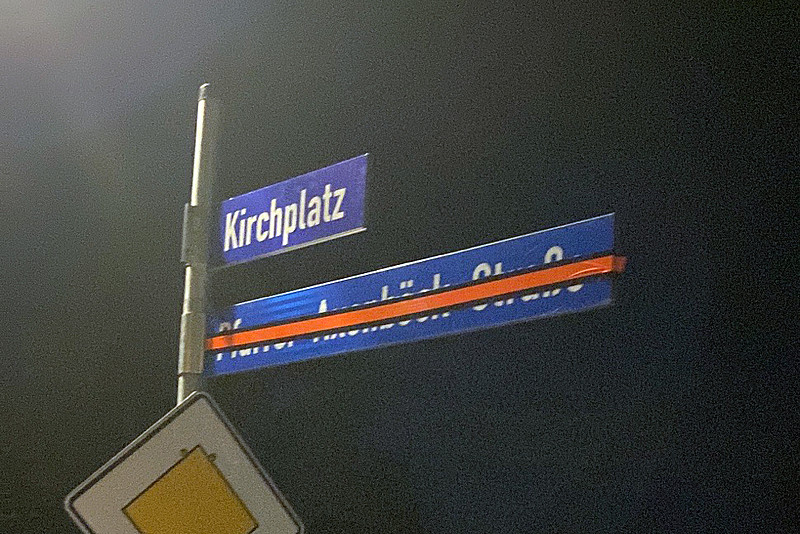 Straßenschild mit der Aufschrift "Kirchplatz", darunter das alte Schild, dessen Name durchgestrichen ist