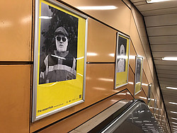 Die eingerahmten schwarz-weiß Portraits auf gelbem Hintergrund hängen neben der Rolltreppe, die zur U-Bahn führt.
