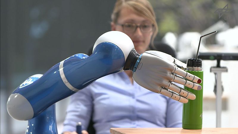 Roboter-Greifarm greift nach Flasche, Frau im Hintergrund