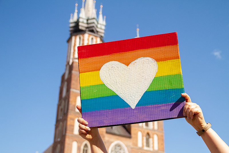 Schild in Regenbogenfarben und mit weißem Herz wird vor Kirchturm in die Höhe gehalten. 