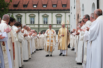 links und rechts stehen viele Priester und durch die Mitte geht der neu geweihte Priester mit Daumen hoch und daneben der Bischof 