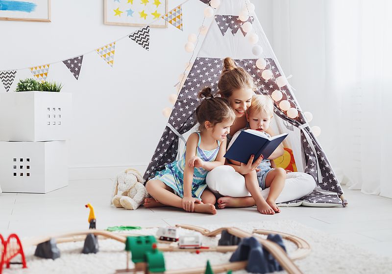 Frau liest zwei Kindern in einem Zelt etwas vor, im Vordergrund Spielzeug