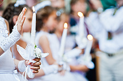 Hand eines Mädchens hält eine Kerze bei der Erstkommunion, im Hintergrund sind weitere Kinder zu sehen
