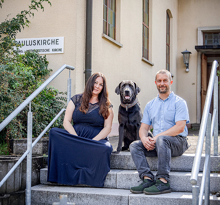 Ellen Weinmann und Florian Düsterwald nebeneinander auf einer Treppe sitzend. In der Mitte zwischen ihnen hockt ein Hund.