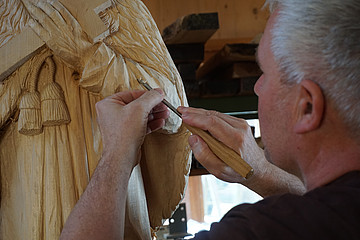 Von hinten seitlich sieht man Christian Dögerl, der mit einem Schnitzeisen Falten des Chorrocks in das Holz der Figur kerbt.
