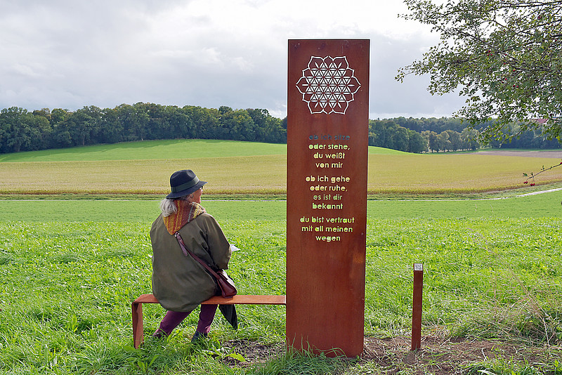 Frau sitzt in grüner Landschaft neben Stele des Ebersberger Meditationswegs