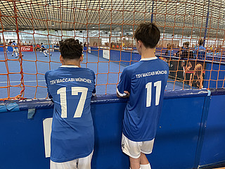 Zwei Buben im blauen Fußballtrikot (weiße Hose) stehen mit dem Rücken zum Betrachter vor einem Tornetz und beobachten ein Fußballspiel.