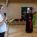 Junge Frau steht mit Mikrofon und beschriebenem Blatt vor Publikum, im Hintergrund erkennbar der Weihbischof.