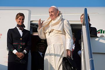 Papst Franziskus steigt aus einem Flugzeug und hebt die Hand zum Gruß, neben ihm zwei Stewardessen. 