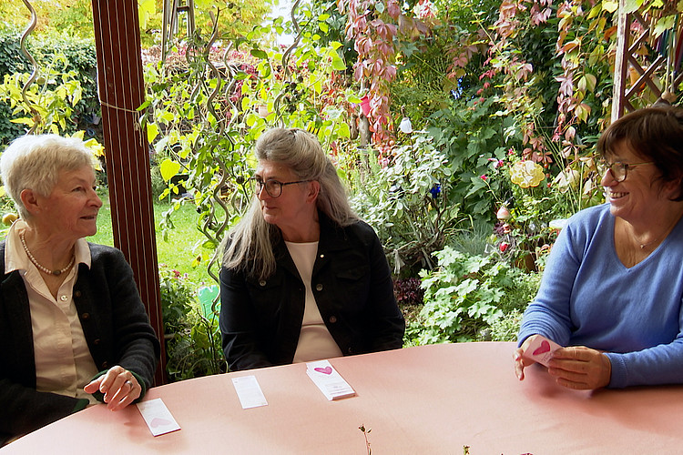 Drei ältere Frauen sitzen um Gartentisch herum und lächeln sich an. 