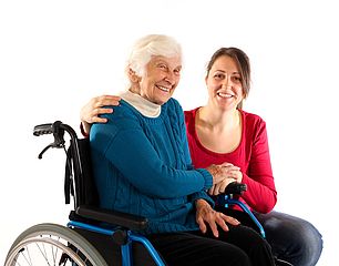Junge Frau kniet neben alter Dame im Rollstuhl.