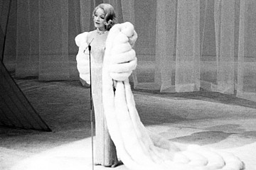 Marlene Dietrich in langem Kleid und Pelzmantel auf der Bühne, vor ihr ein Mikrofon, hinter ihr ein geschlossener Vorhang