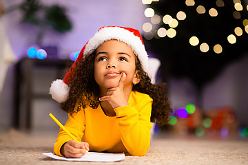 Kleines Mädchen mit Weihnachtsmannmütze liegt auf dem Boden vor einem Zettel, hält einen Stift in der Hand und schaut nach oben, während es sich einen Finger an die Wange hält. 