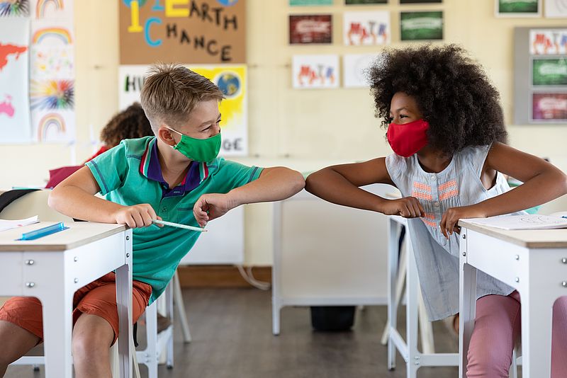 Zwei Kinder mit Maske grüßen sich mit Ellbogen im Klassenzimmer