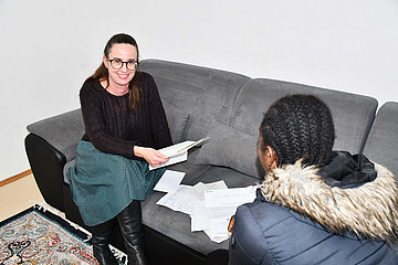 Zwei Frauen sitzen auf einem grauen Sofa, zwischen ihnen liegen Dokumente.